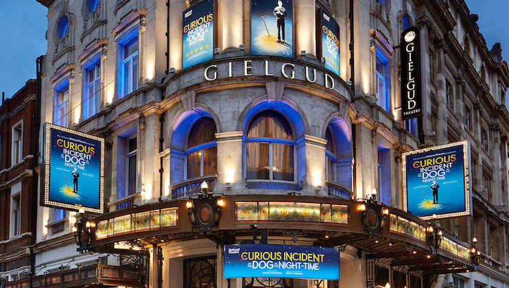 The facade of the Gielgud Theatre, adorned with multiple banners advertising The Curious Incident of the Dog in the Night-Time. The building is illuminated at night, showcasing its ornate architecture and brightly lit signage.