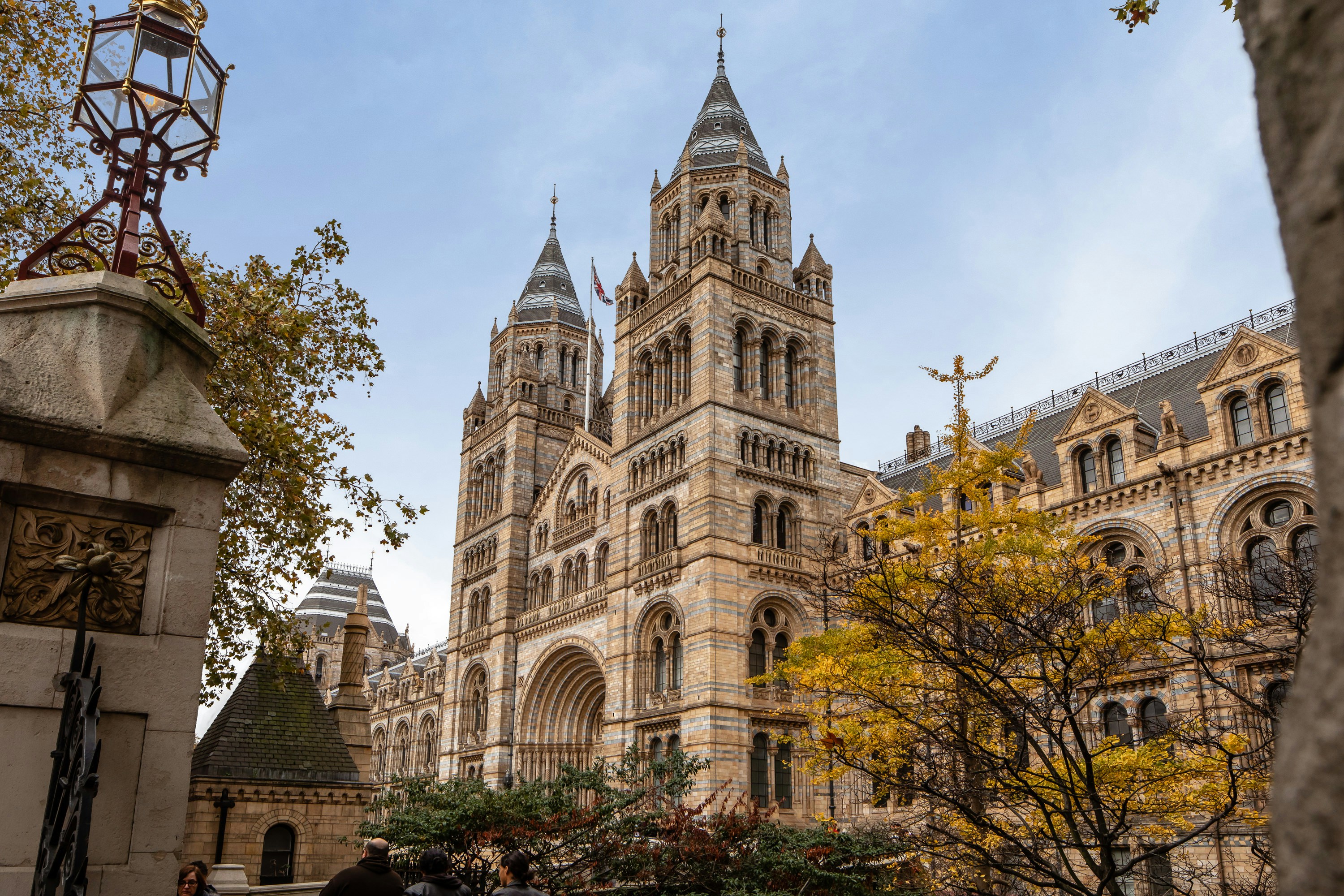 A large, ornate Gothic Revival building with two prominent towers stands under a partly cloudy sky. The facade is detailed with arches and stonework. Trees with autumn leaves are in the foreground, and parts of a surrounding structure are visible.