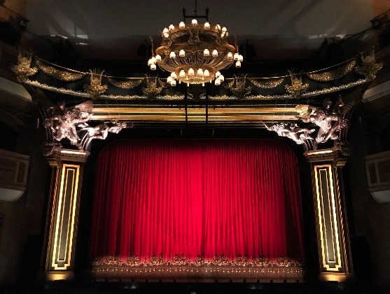 A grand theater stage with a closed red curtain, framed by ornate gold decorations and statues on either side. A large, elegant chandelier hangs from the center of the ceiling above the stage. The ambiance is opulent and classic.