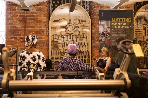 Three people are engaged in conversation in front of a wall display featuring an array of vintage tools. One woman, in work attire, explains something to two others, who are wearing colorful, patterned headscarves. The setting is inside an industrial-themed museum.