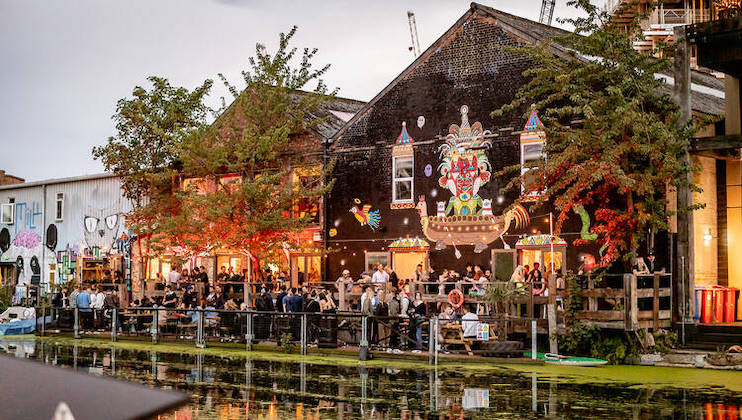 A vibrant riverside scene with a crowd of people outside a lively building featuring colorful, whimsical street art on its exterior. The building, adorned with festive lights, sits by a calm river with lush greenery and reflections in the water, creating a festive atmosphere.