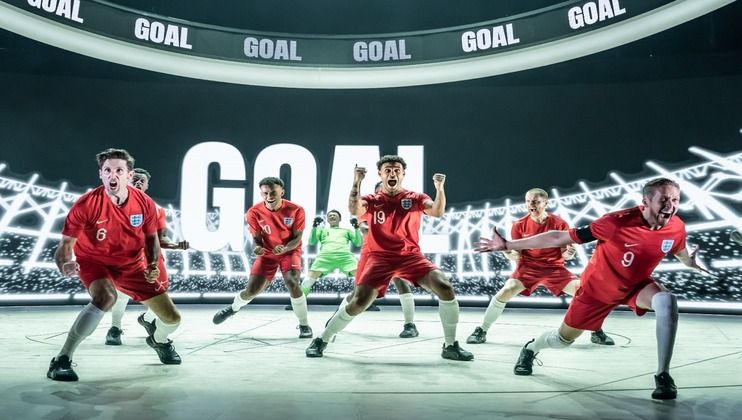 A group of soccer players in red uniforms celebrate energetically in front of a large screen displaying the word GOAL in bold letters. The background features illuminated stadium lights, capturing the intense atmosphere of the moment.