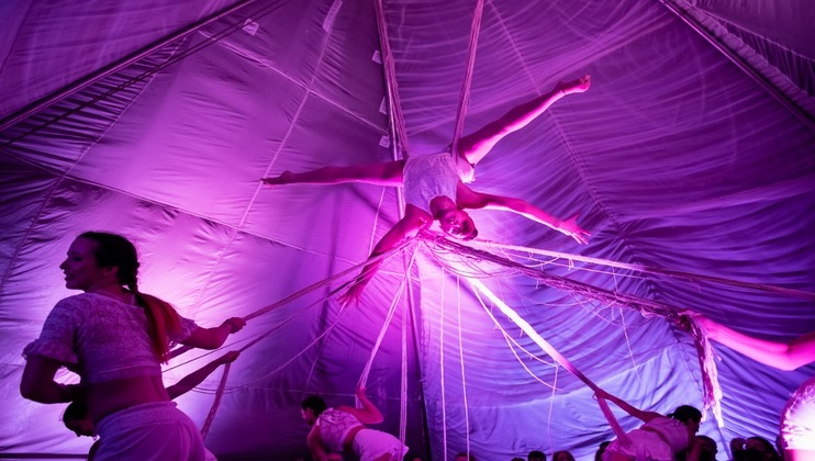 Aerial acrobats are performing inside a tent illuminated with purple and pink lights. One acrobat is in the air, supported by ropes, legs outstretched in a split. Several other acrobats in similar attire are on the ground, interacting with the ropes.