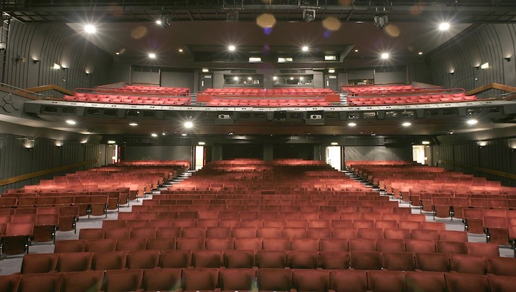 An empty auditorium features rows of red cushioned seats, a balcony section filled with additional seating above, and a low-lit stage area at the front. The space is well-lit with overhead lighting, highlighting the clean and orderly arrangement of the theater.