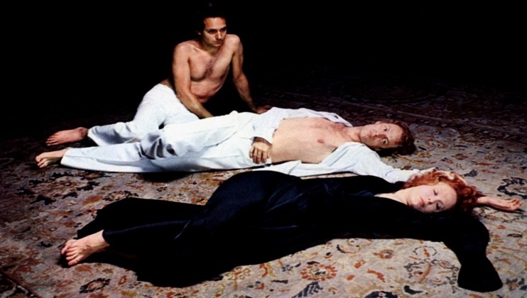 Three individuals are lying on an ornately patterned carpet. A shirtless person in white pants is sitting up in the background. In the foreground, a shirtless person with red hair in white pants and another person with short hair in a black outfit lie on their backs.