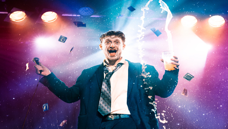 A man in a suit joyfully holds a microphone in one hand and a drink in the other, with the drink splashing upward. Multicolored lights and confetti dramatically illuminate the background, indicating a lively, celebratory atmosphere.