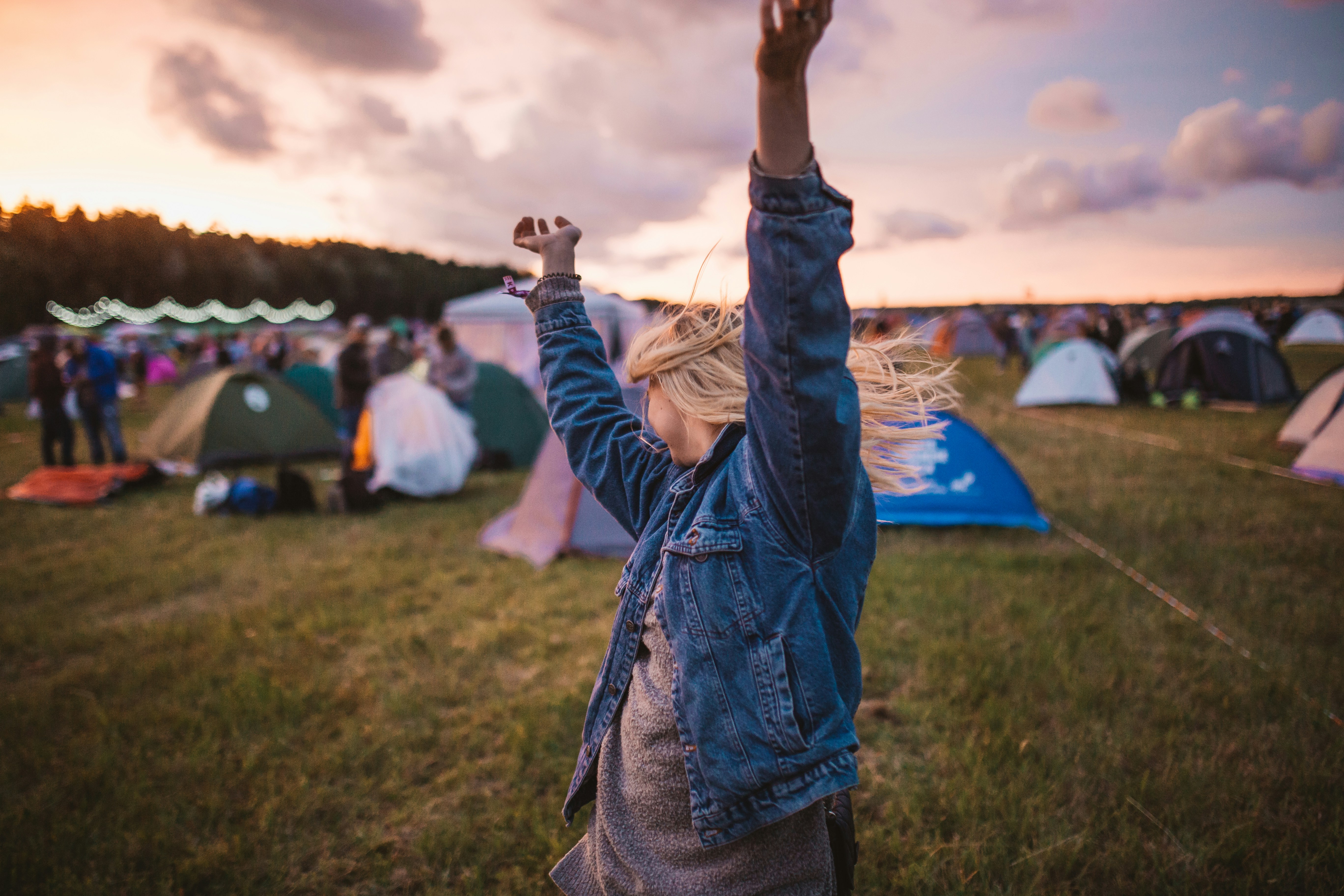 A person with blonde hair wearing a denim jacket dances with their arms raised in a grassy field, surrounded by tents. The sun is setting and the sky is filled with clouds, creating a warm, vibrant atmosphere.