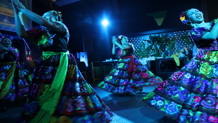 A group of women dressed in vibrant, traditional Mexican clothing are dancing joyfully under blue lights. They wear colorful dresses adorned with floral patterns and ribbons, and their expressions show delight and celebration. The background features festive decorations.