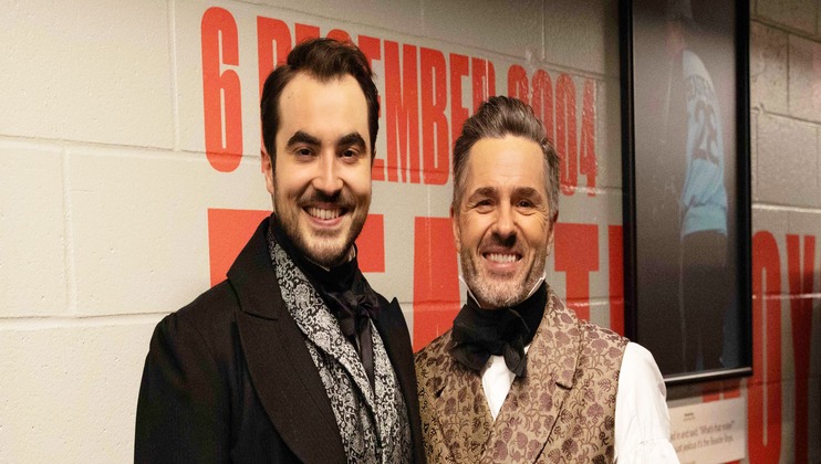 Two men are dressed in period costumes, likely from the Victorian era. One wears a dark suit with a high collar and cravat, while the other dons a patterned waistcoat and bow tie. They are standing against a wall with partially visible red text and framed pictures.