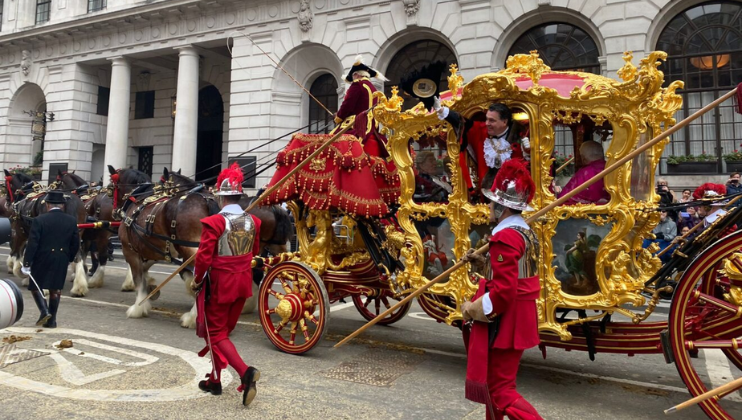 A lavish, gold-adorned carriage is being pulled by horses through a city street during a parade. People in red and gold ornate costumes escort the carriage. The surrounding architecture includes grand, arched windows and columns. Onlookers are visible in the background.