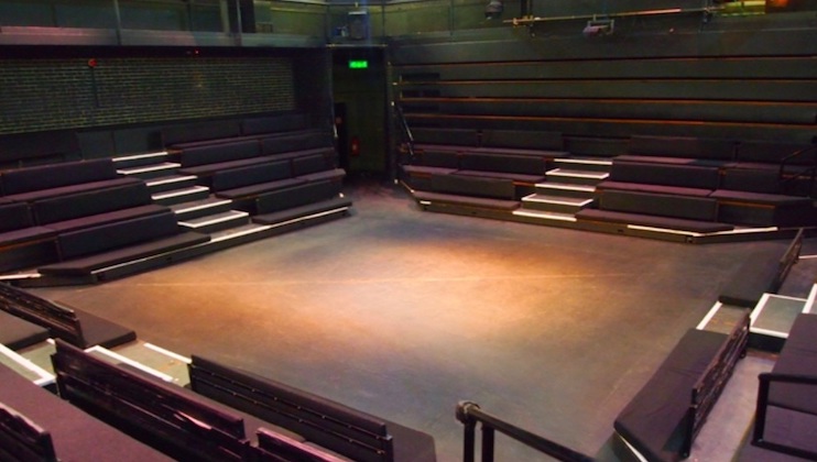 A modern, empty theater with a black box style stage. The seating arrangement creates an intimate atmosphere, with tiered seats on three sides of the stage. The lighting is dim, casting a soft glow on the center of the stage area.