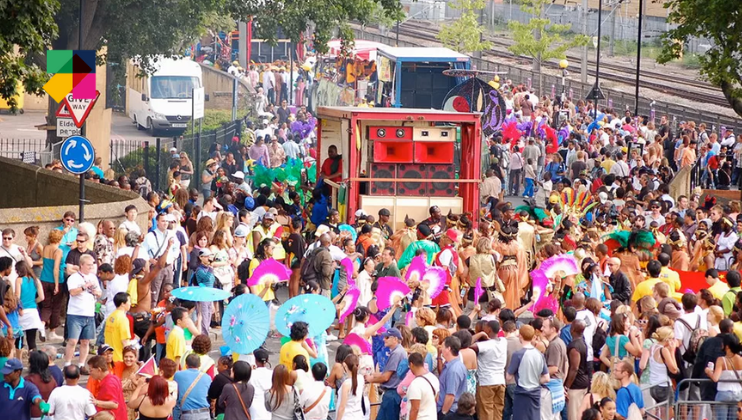 A vibrant street scene at a festival with diverse crowds. People are wearing colorful costumes and accessories, such as feathers and umbrellas. A red parade float is in the center. The background shows trees, vehicles, and a railway track.