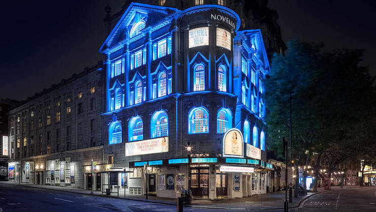 A nighttime view of the Novello Theatre in London, beautifully illuminated with blue lights. The building's historic architecture is highlighted, and bright marquees display show information. The street in front is quiet and nearly empty.