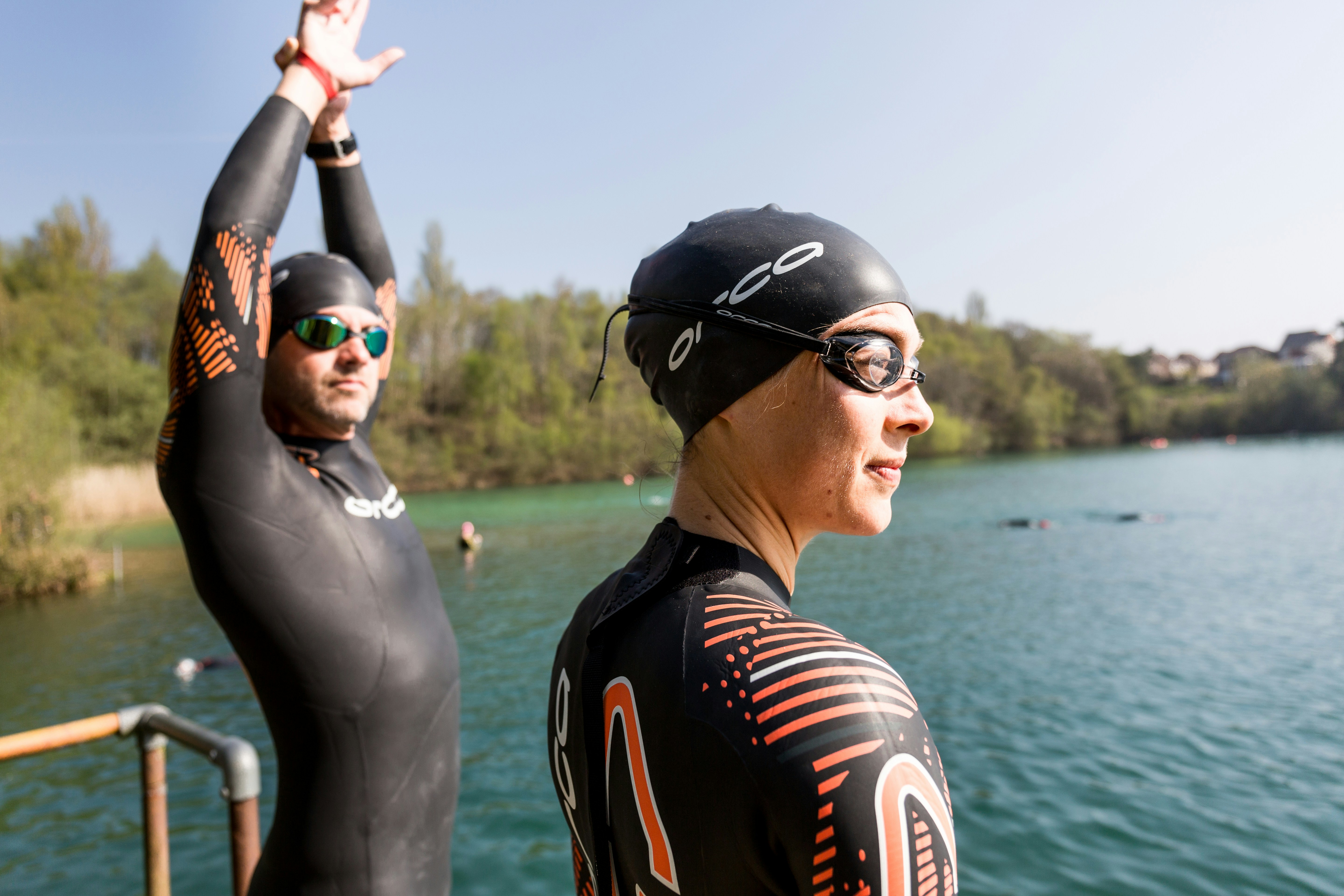 Two swimmers in wetsuits and swim caps stand by a serene lake, preparing for an open water swim. One person stretches with arms raised, while the other looks out over the water. The background features trees and a clear sky.