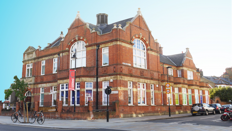 A large red brick building with arched windows and ornate detailing, featuring a bookshop banner on the side. Bicycles are parked near the entrance, and several cars are parked along the street. The sky is clear and blue, and the street is empty.