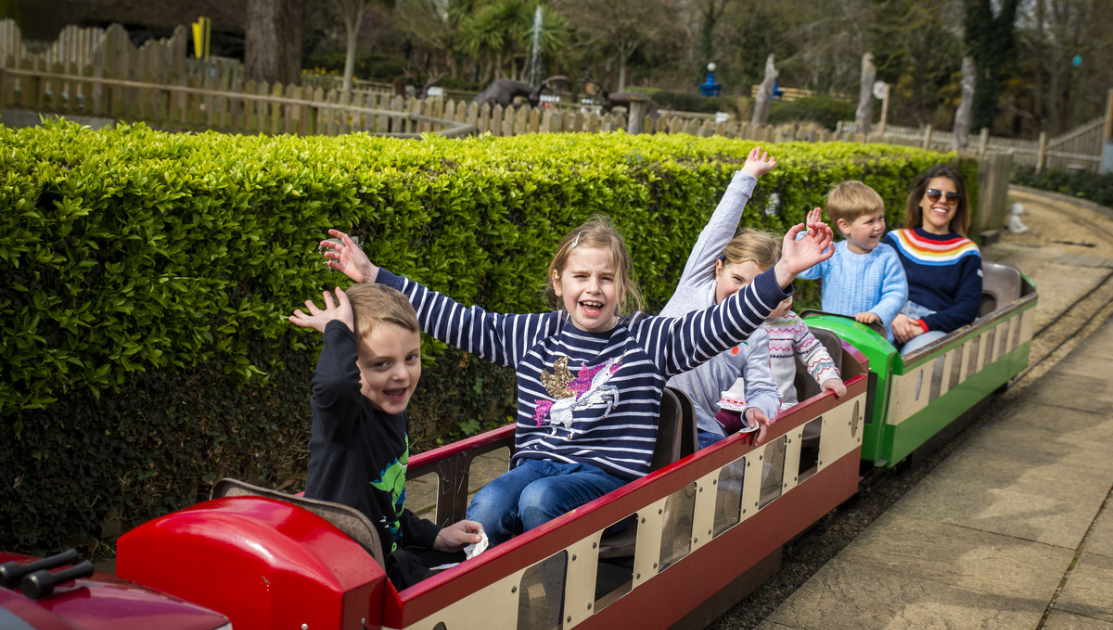 A group of children and an adult ride on a red and green miniature train at an amusement park. The children, sitting in open-air cars, wave their hands in excitement with wide smiles. The track is surrounded by greenery and a wooden fence, under a clear sky.