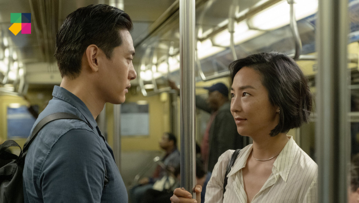 A man and woman stand facing each other on a subway, holding onto a shared pole. The woman, with short black hair and a white blouse, looks at the man, who has short dark hair and wears a blue shirt. Other passengers are seated and standing in the background.