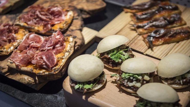 An assortment of gourmet appetizers including cured meat on toasted bread, small sandwiches with greens and meat, and slices of bread topped with anchovies are arranged on wooden platters. The dishes are displayed on a dark table.