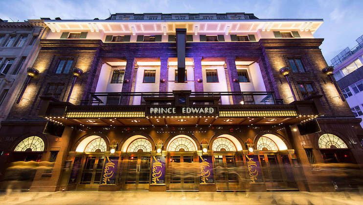 A nighttime view of the Prince Edward Theatre, an elegant building with an illuminated marquee and arched windows. The facade is decorated with columns and brickwork, while the marquee casts a warm glow, highlighting the name Prince Edward in white letters.