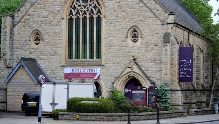 A stone building with arched windows and a large, pointed entrance houses the Putney Arts Theatre. A banner above the entrance advertises a production of West Side Story. A black van and a white truck are parked in front, along with some greenery.