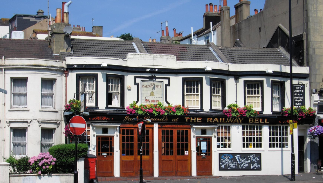 A quaint pub called The Railway Bell has a white and black facade with red brick accents. Vibrant flower boxes adorn the windows. There are multiple chimneys on the roof. A No Entry sign is in the foreground next to chalkboard signs promoting events.