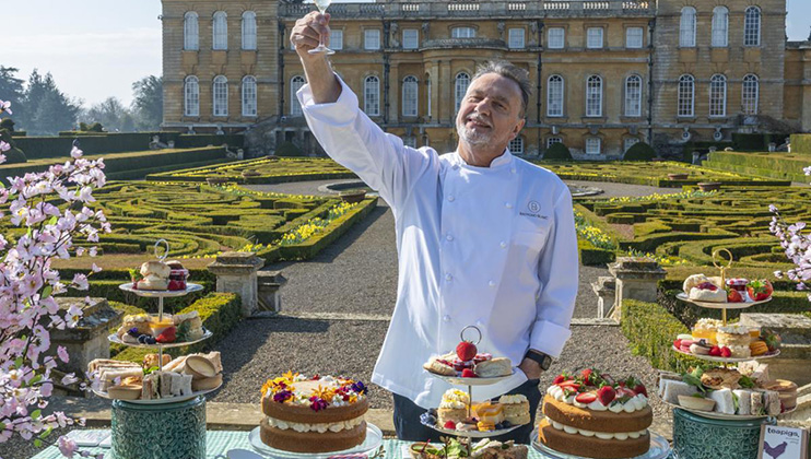 A smiling chef in a white uniform lifts a drink amidst a garden setting with an ornate building in the background. Several tables display an assortment of colorful pastries, cakes, and sandwiches on tiered stands, surrounded by blooming pink flowers.