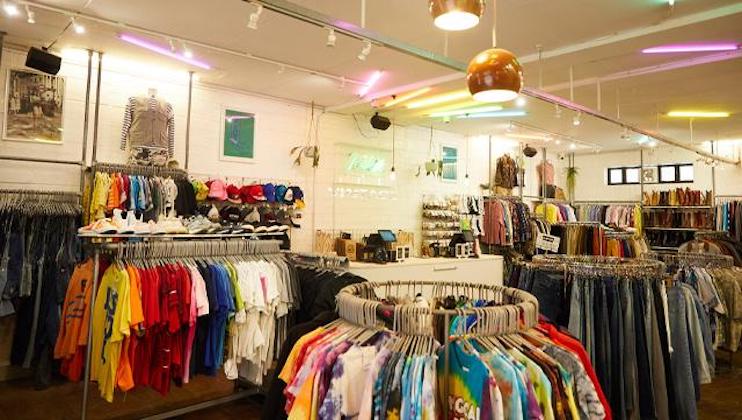 A vibrant clothing store with colorful neon lights on the ceiling. The store features various racks of clothes including T-shirts, jackets, and jeans. A central circular rack displays tie-dye shirts. Accessories and hats are visible on shelves in the background.