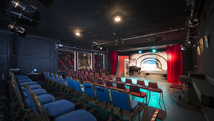 A small theater with a stage decorated with red curtains and a brick wall backdrop. The stage is lit with blue and white lights. Rows of blue and red chairs are arranged for the audience in the foreground. The theater has a cozy and intimate atmosphere.