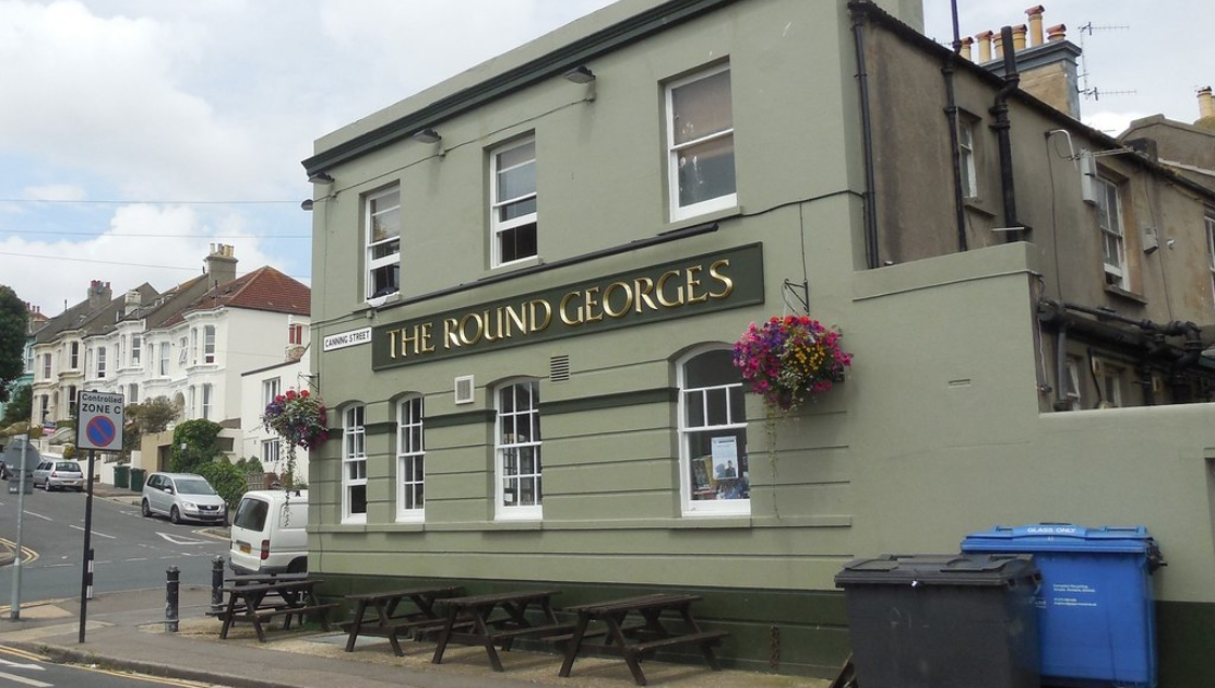 A two-story pub with olive green exterior walls and a sign reading The Round Georges in gold letters. Flower baskets hang near the entrance. Wooden picnic tables are positioned outside. Residential homes and parked cars line the street beside the pub.