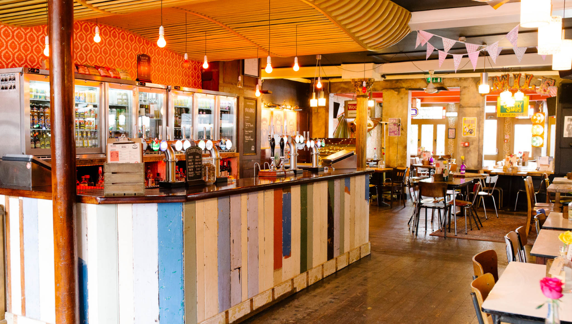 A brightly lit, eclectic bar and restaurant with a mix of rustic and modern decor. The bar is adorned with colorful, weathered wood panels. Pendant lights hang from a wave-patterned ceiling. Tables and chairs are scattered throughout the space with various decorations.