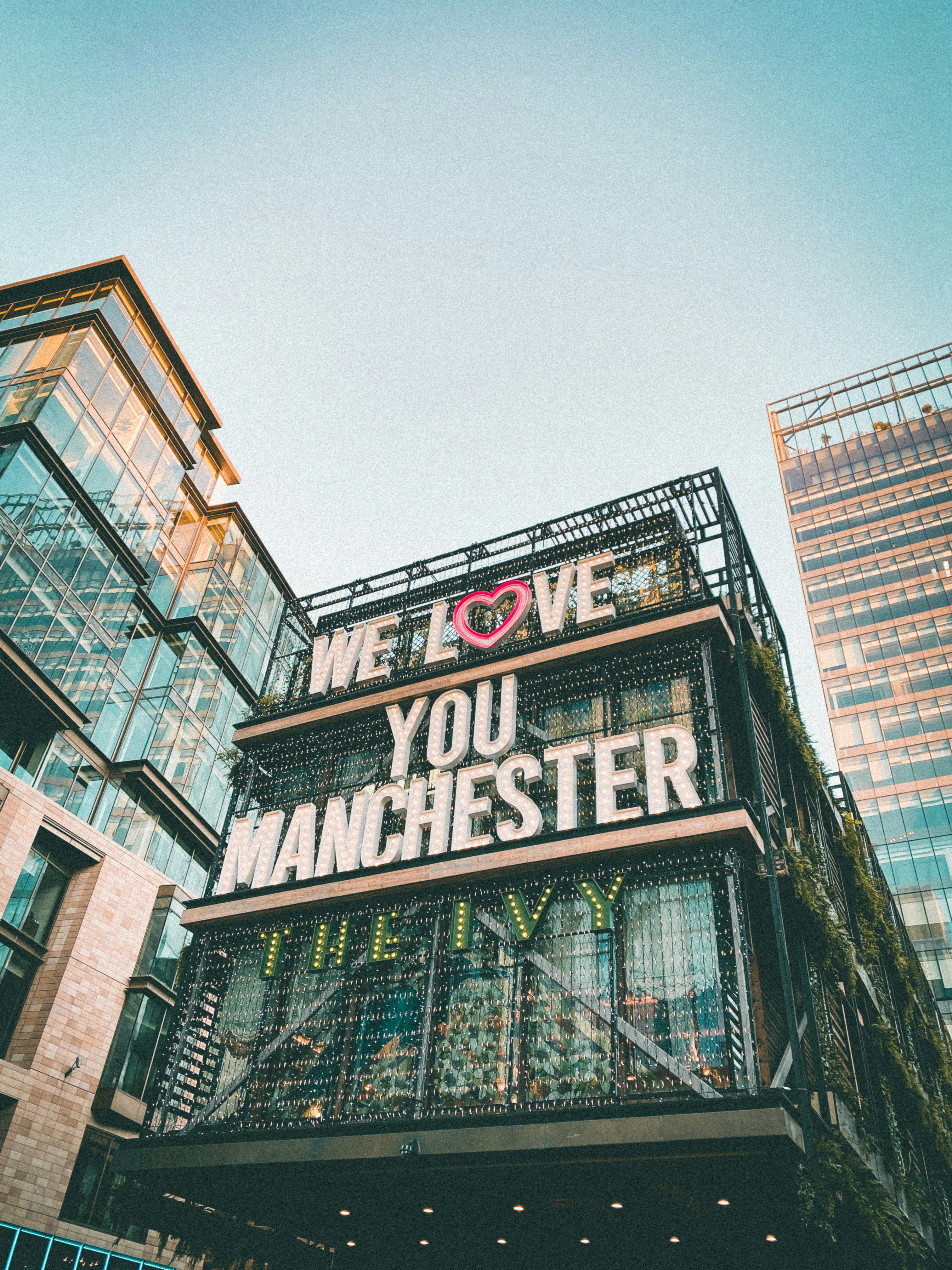 A modern urban scene featuring a building adorned with large neon text that reads WE LOVE YOU MANCHESTER with a pink heart symbol. Surrounding the building are glass-fronted high-rises against a clear sky.