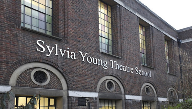 Exterior of a brick building with arched windows and a sign reading Sylvia Young Theatre School in white letters. The building has several rectangular stained glass windows above the arches and appears to have an upper floor.