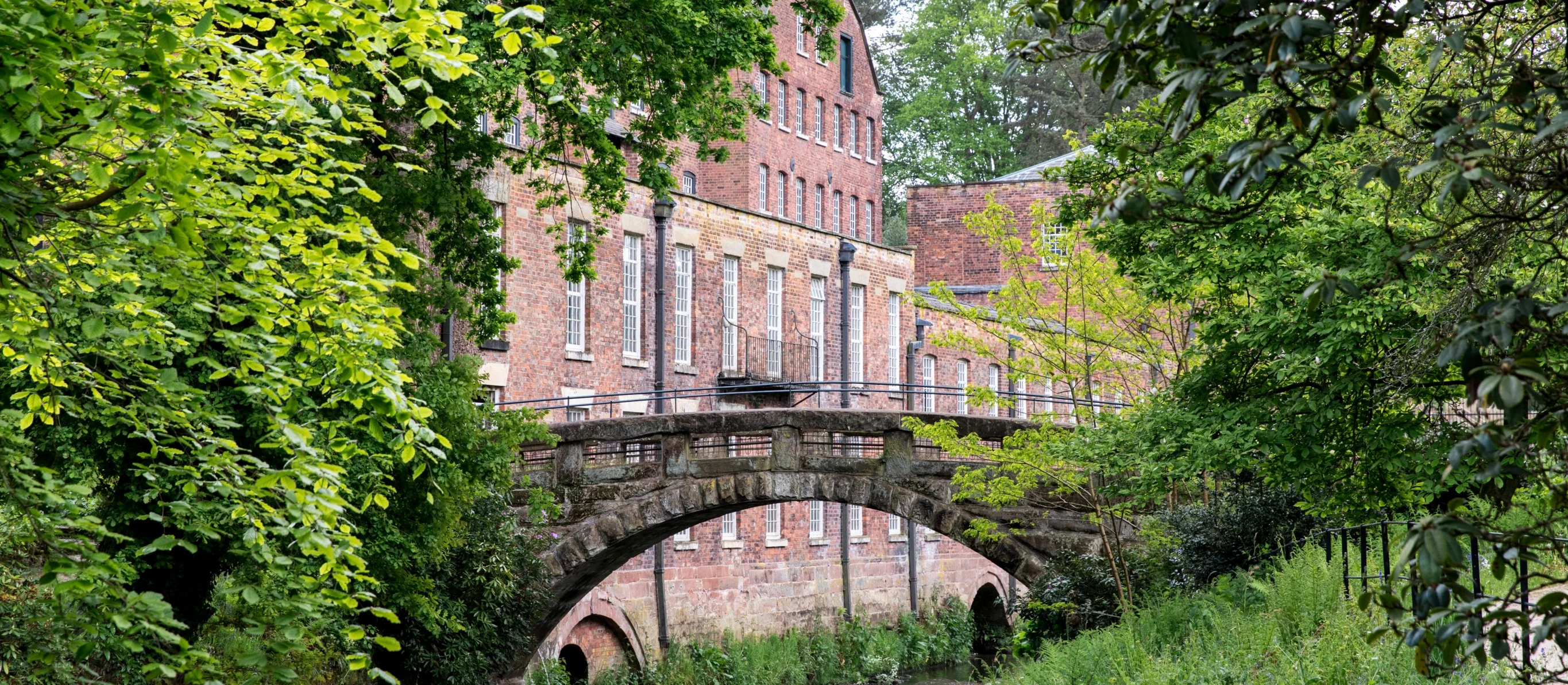 A serene brick building, partially obscured by lush green foliage, stands alongside a stone arch bridge over a narrow waterway. The scene is quiet and picturesque, emphasizing the natural and historic elements harmoniously coexisting.