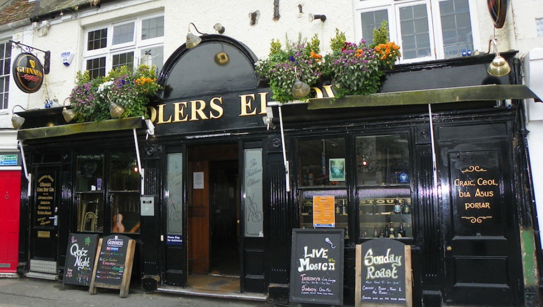 A traditional pub with a black façade, named Solers Elbow. It has hanging lanterns, flower boxes with vibrant flowers above the entrance, and several chalkboard signs advertising a Sunday roast, live music, and quiz night. The windows have white frames.