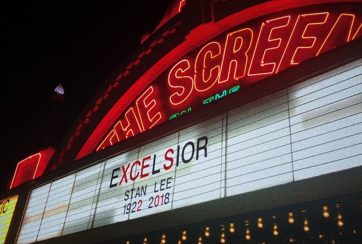 A theater marquee illuminated by red neon lights reads EXCELSIOR STAN LEE 1922-2018. The text is displayed beneath the word SCREEN in large red letters. The image appears to be a night scene, with the lights providing a bright contrast against the dark sky.