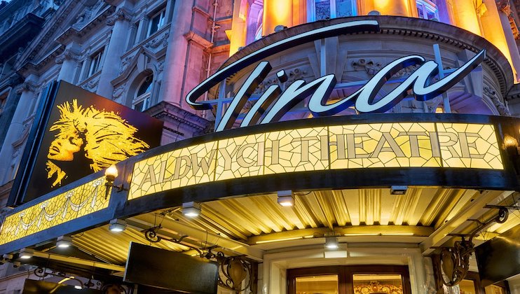 The image shows the entrance to the Aldwych Theatre adorned with signage for the musical Tina. The theater's marquee is illuminated, with the show's logo, a stylized portrait of Tina Turner, prominently displayed above. The building's ornate architecture is visible.