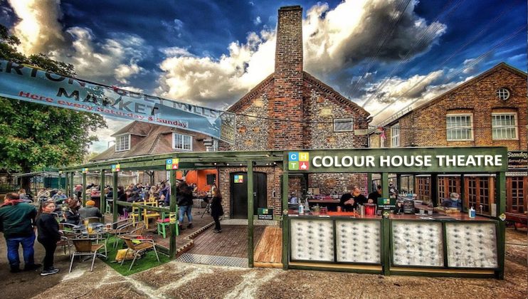 Vibrant scene at the Colour House Theatre with a colorful building and outdoor seating. People gather in the market area under a banner. The sky above is partly cloudy, adding a dramatic backdrop to the lively setting.