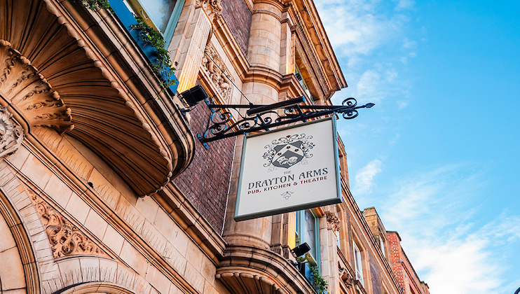 A sign reading Drayton Arms Pub, Kitchen & Theatre hangs from an ornate bracket on the exterior of a historic brick building under a blue sky.