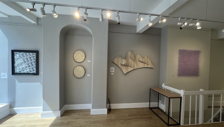 A modern art gallery with neutral-toned walls displaying four pieces of art: a textured white and black frame, two circular pieces with intricate patterns, an abstract wooden sculpture, and a large purple textured canvas. The room has wooden flooring and track lighting.