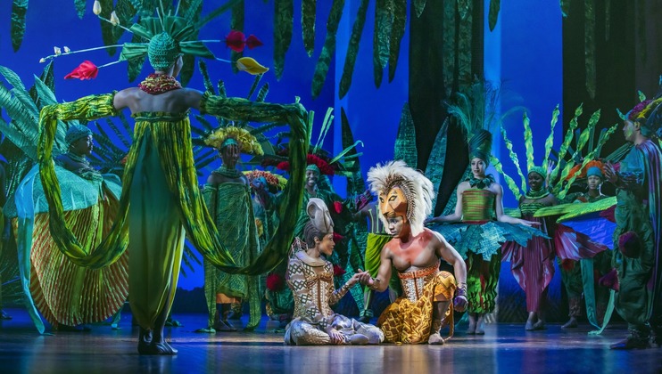 A vibrant stage performance scene from a musical, featuring actors in elaborate costumes. Center stage, two characters sit, while others stand around them in colorful headdresses and leafy attire, creating a lively jungle atmosphere. The background is blue with leafy decorations.