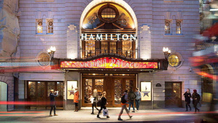 The entrance to Victoria Palace Theatre in London at night. The marquee reads “Hamilton,” brightly lit above the doors. People are walking and standing in front of the theater. The building is illuminated, creating a vibrant and lively atmosphere.