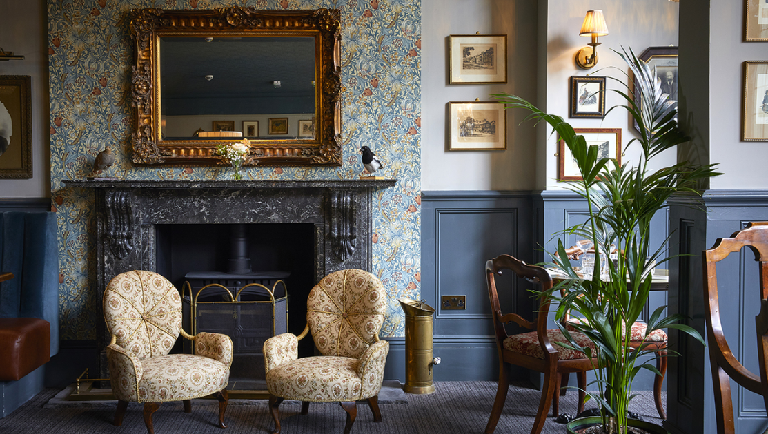 A cozy room features two patterned armchairs in front of a dark stone fireplace with an ornate gold mirror above it. Wallpaper with floral designs decorates the upper walls, while framed pictures and a tall potted plant add to the elegant, vintage ambiance.