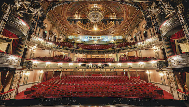 A grand theater auditorium with ornate architectural details, featuring luxurious red seating on the main floor and two balconies. The ceiling is adorned with an intricate chandelier, and the stage is visible at the front. The atmosphere is elegant and historic.