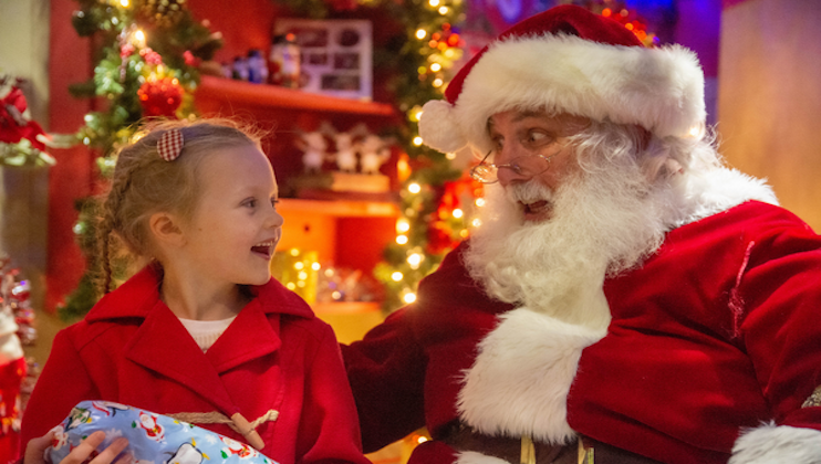 A young girl in a red coat smiles at a man dressed as Santa Claus. Santa, in his red suit and hat with white trim, looks at her with a joyful expression. The background is festive with Christmas lights, decorations, and a tree. The girl holds a wrapped gift.