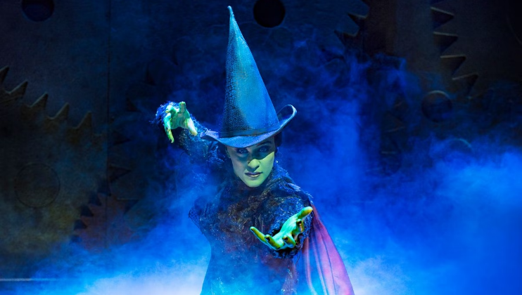 A person with green skin wearing a tall, pointed black hat and a dark costume is dramatically posing with outstretched hands. Blue fog surrounds them, creating a mystical atmosphere. The background shows large gears and mechanical elements.