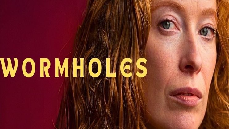 A close-up of a person with red hair and light eyes looks pensively at the camera against a maroon background. The word WORMHOLES is written in large yellow letters to the left of the person's face.