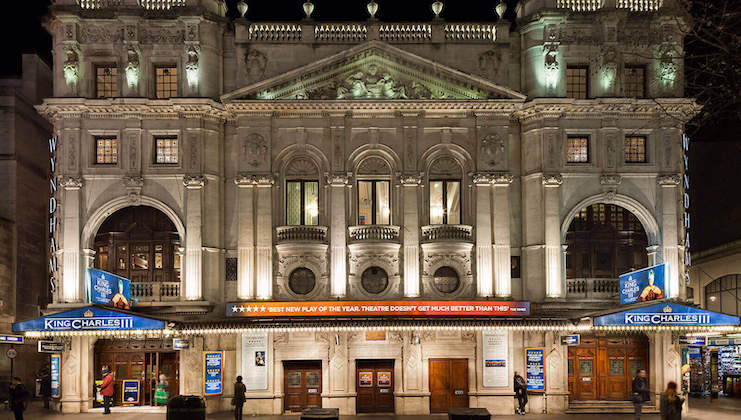 A grand, illuminated theater front featuring a neoclassical architectural design is shown at night. There are intricate sculptures above the large arched windows, and a marquee displays the play King Charles III. People are seen walking by the entrance.